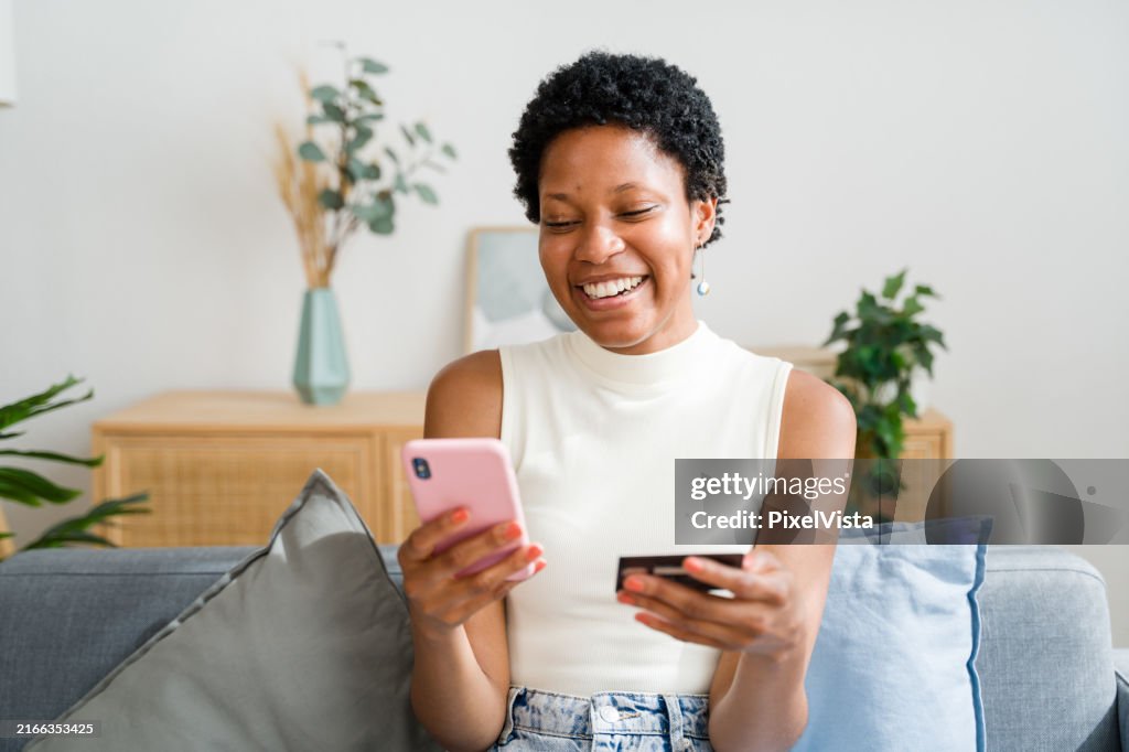 Young black woman sitting on sofa is using smartphone for online shopping with credit card