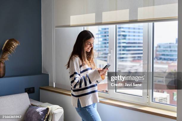 happy woman is using a smartphone while standing by the window in a modern living room - rapariga no quarto tecnologia imagens e fotografias de stock