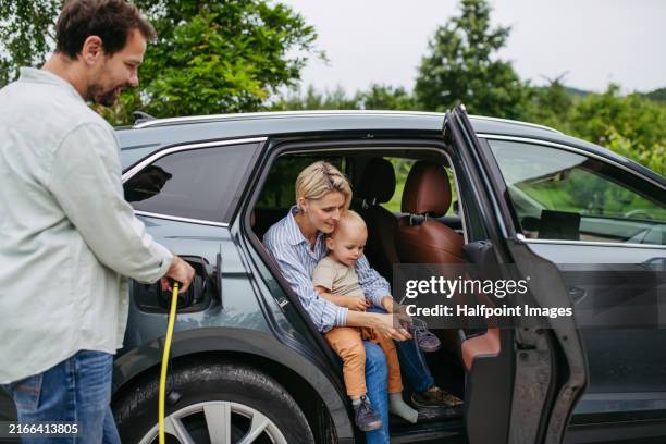 family and electric car. father plugging charger in charging port, mother and toddler getting out of car. - hybrid car stock pictures, royalty-free photos & images