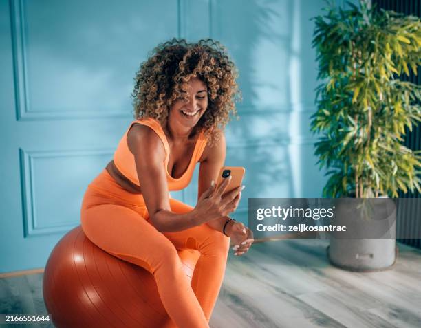 young sporty woman sitting on fitness ball in living room and using a phone - sportkleding stockfoto's en -beelden