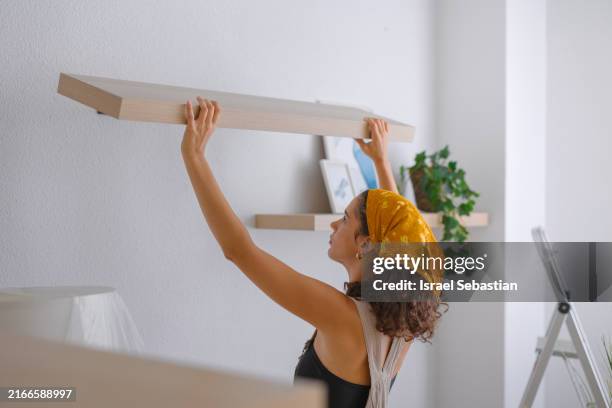 young woman installing wooden shelf on white wall - scaffale foto e immagini stock
