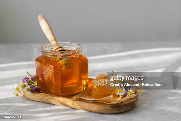 honey jar, honeycomb and wild flowers. fresh organic honey in glass jar on white table background with shadow sunlight. - honeycomb toffee stock pictures, royalty-free photos & images