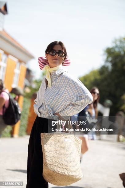 Guest wears black pants, blue and white stripe shirt, beige bag, white and red hair bow and pale yellow neck scarf outside the Skall Studio show...