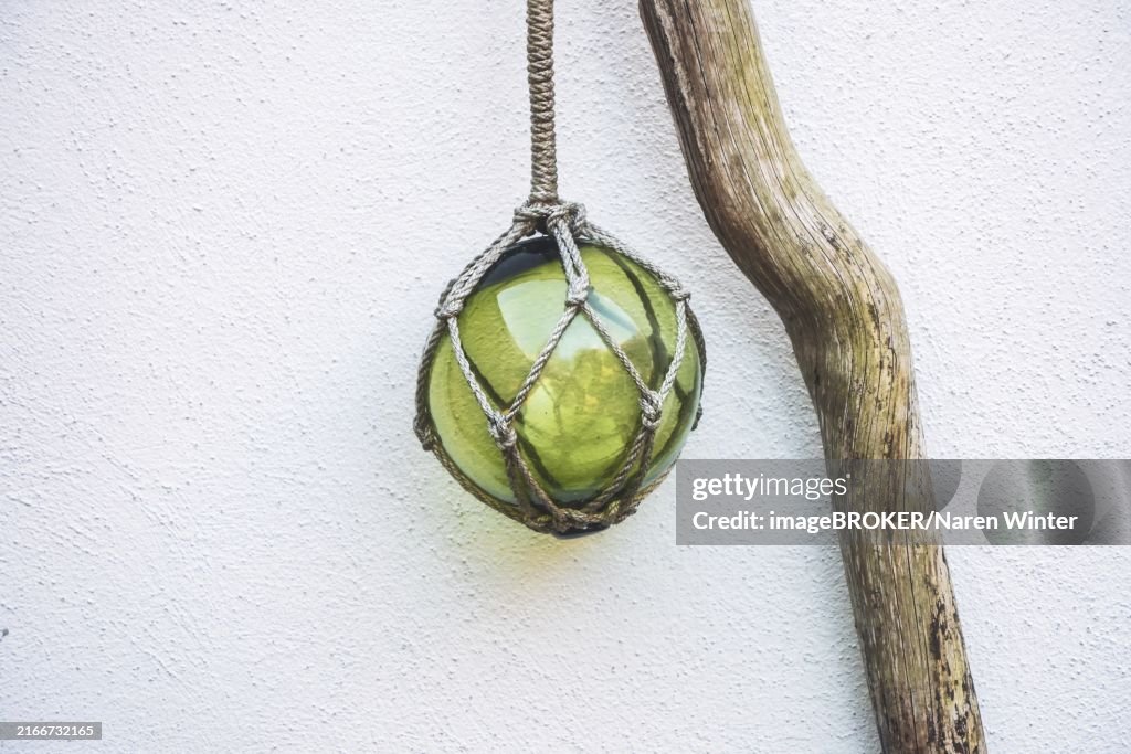 Glass float hanging as decoration on a driftwood against a white wall, the historic item was used to keep the fishing nets afloat, copy space, selected focus