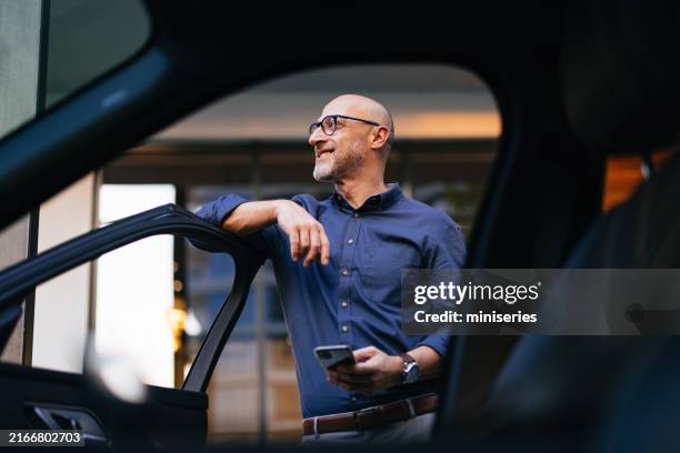 confident businessman standing by car with smartphone in city - carro imagens e fotografias de stock