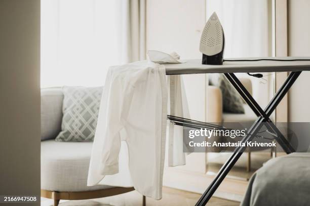 electric iron and white shirt on ironing board in luxury interior. - repassage photos et images de collection
