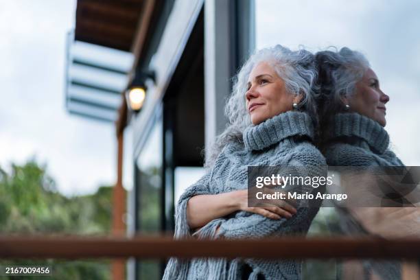 mujer mira a lo lejos en un retrato mientras se apoya en la ventana en una hermosa mañana - adulto de mediana edad fotografías e imágenes de stock