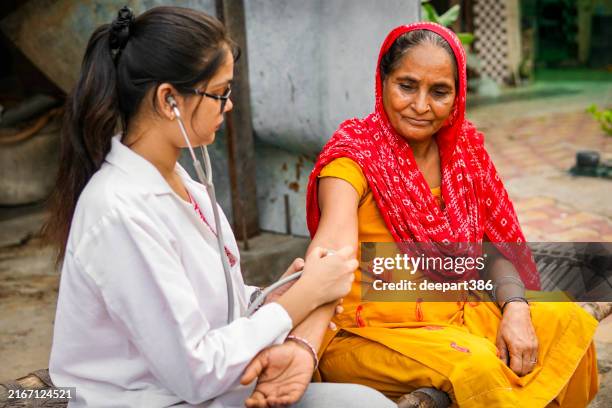 female indian doctor examining a rural senior woman patient with stethoscope during house visit in village. - developing countries stock pictures, royalty-free photos & images