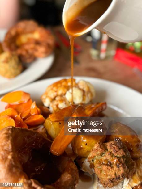 image en gros plan du pudding géant du yorkshire recouvert de sauce sur une assiette blanche contenant le repas du dîner rôti du dimanche, poulet rôti, carottes, chou-fleur, panais, pommes de terre, boule de farce, personne méconnaissable versant la s - accompagnement photos et images de collection