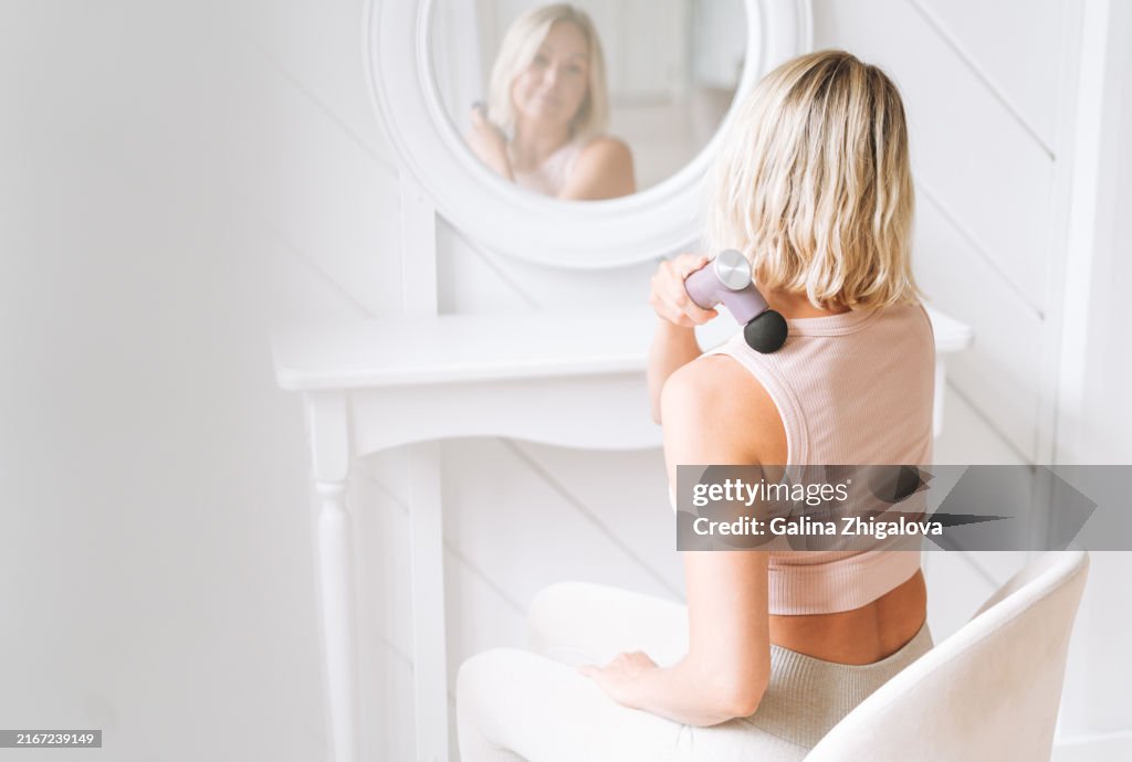 Beautiful blonde woman giving herself neck and collar area massage using percussion massager in front of mirror at home