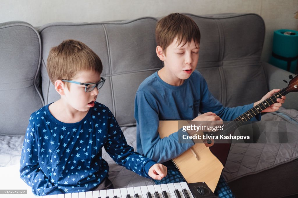 Two boys 10 and 12 years old play the balalaika and synthesizer at home in pajamas while sitting on the couch