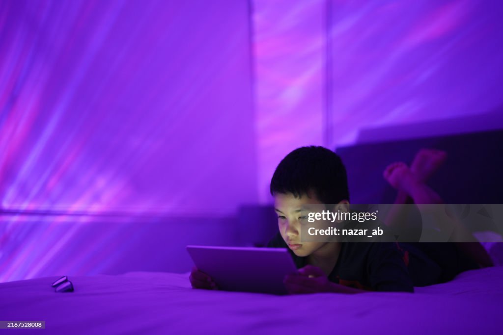 Boy lying on bed using a tablet with a glowing purple light in a dark room creating a magical ambiance.