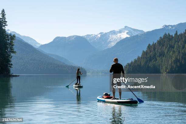 tabuleiros de sup de remo de casal no lago da montanha, whistler - stand up paddle boarding lake - fotografias e filmes do acervo
