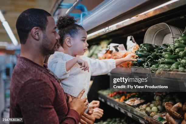 father holds his daughter while picking out vegetables at the store - zwart etniciteit stockfoto's en -beelden