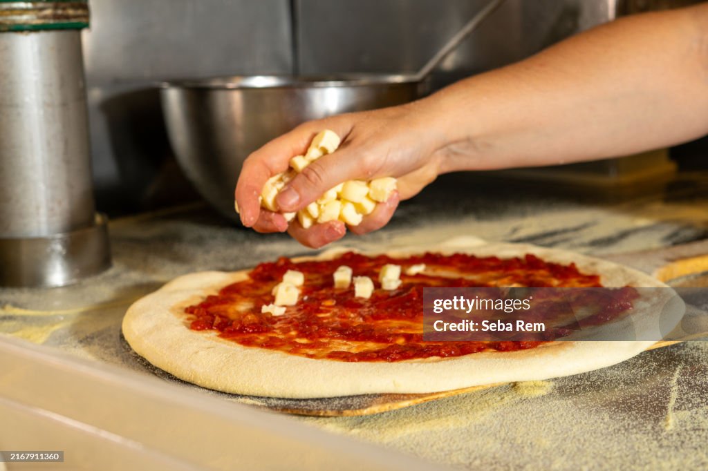 Hand of a professional chef placing fresh mozzarella cubes on pizza dough with tomato sauce, preparing an artisanal pizza in a restaurant kitchen.