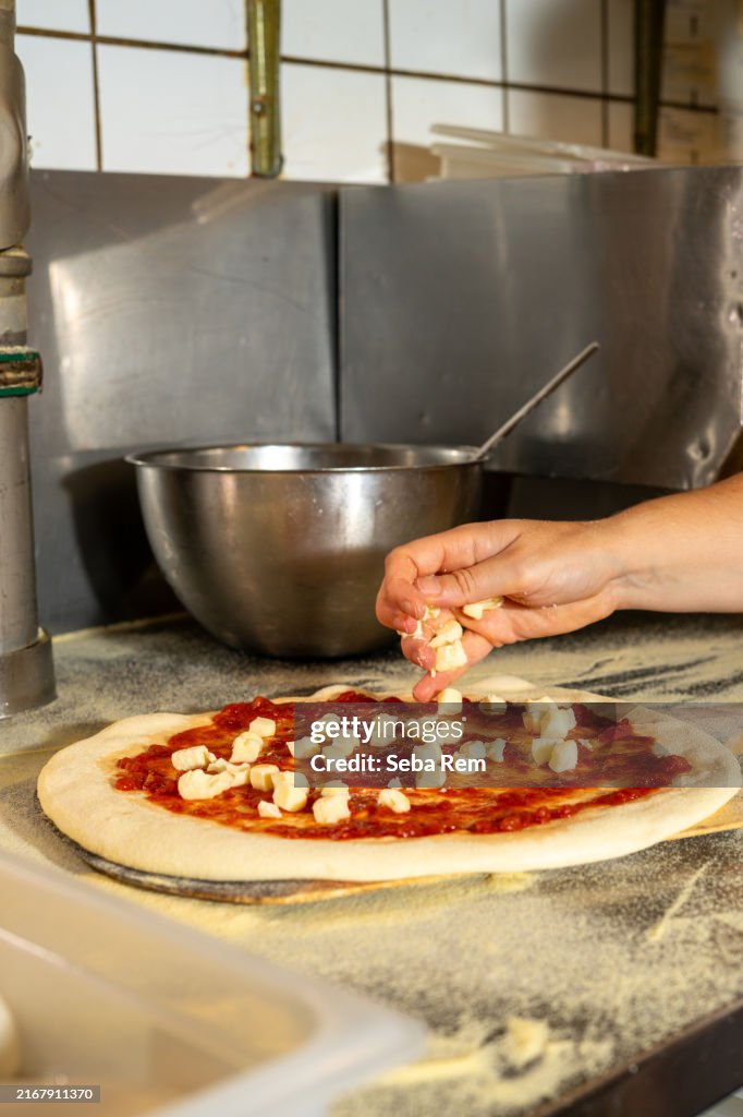 Close-up of a chef's hand sprinkling mozzarella cheese on pizza dough with tomato sauce in a professional pizzeria kitchen, ready for baking.