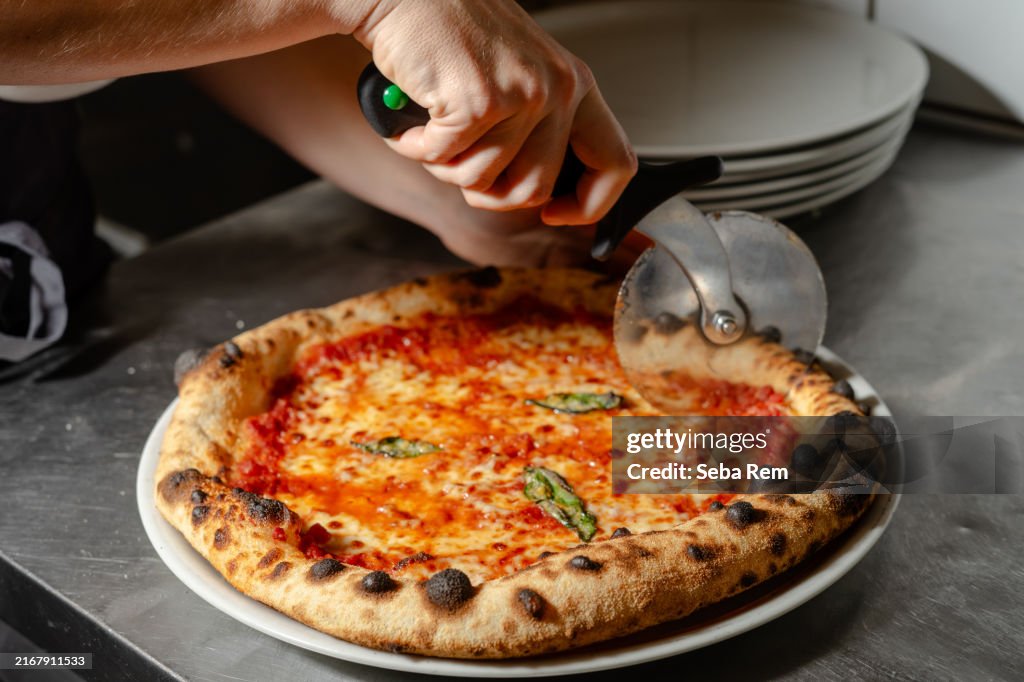 Close-up of a freshly baked Margherita pizza being sliced with a pizza cutter in a commercial kitchen. Crispy crust, melted cheese, and basil leaves on a traditional Italian pizza.