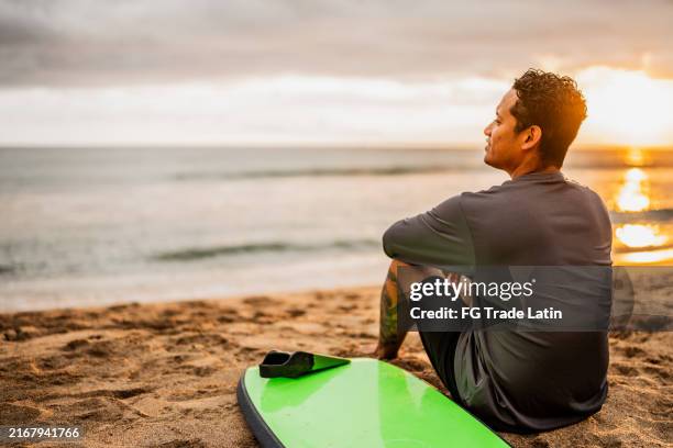 mid adult surfer man contemplating while looking away on the beach - surfer stock pictures, royalty-free photos & images