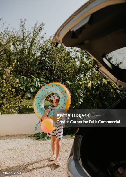 a child walking towards a car carrying a colourful donut pool float and a beach ball on a sunny day in a garden setting - the trunk of the car is open, as she prepares to pack the boot full of pool inflatables - car cover in driveway stock pictures, royalty-free photos & images