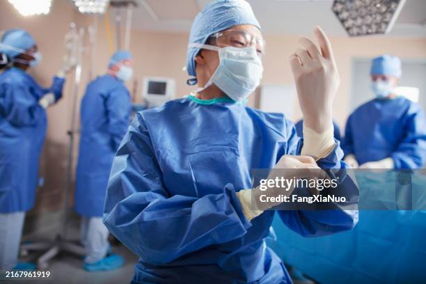 asian chinese mid adult man doctor surgeon nurse puts on surgical gloves in operating room in hospital with medical team - operatiegewaad stockfoto's en -beelden
