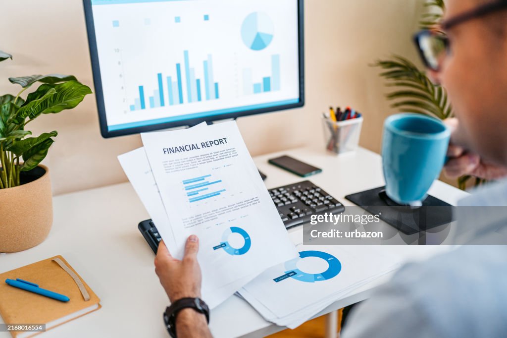 Mid-Adult Man Going Over Paper Documents While Working On A Computer At The Office