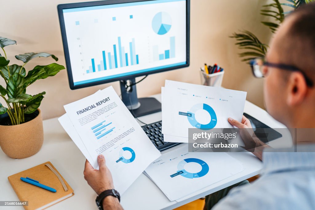 Mid-Adult Man Going Over Paper Documents While Working On A Computer At The Office