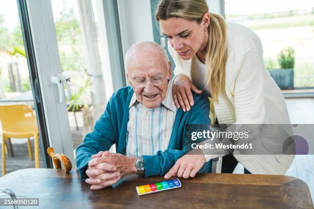woman explaining medicines in multi colored pill box to senior man sitting at table - tablettenbox deutsch stock-fotos und bilder