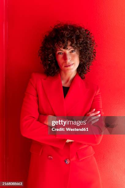 confident businesswoman standing with arms crossed near red wall - roter anzug stock-fotos und bilder