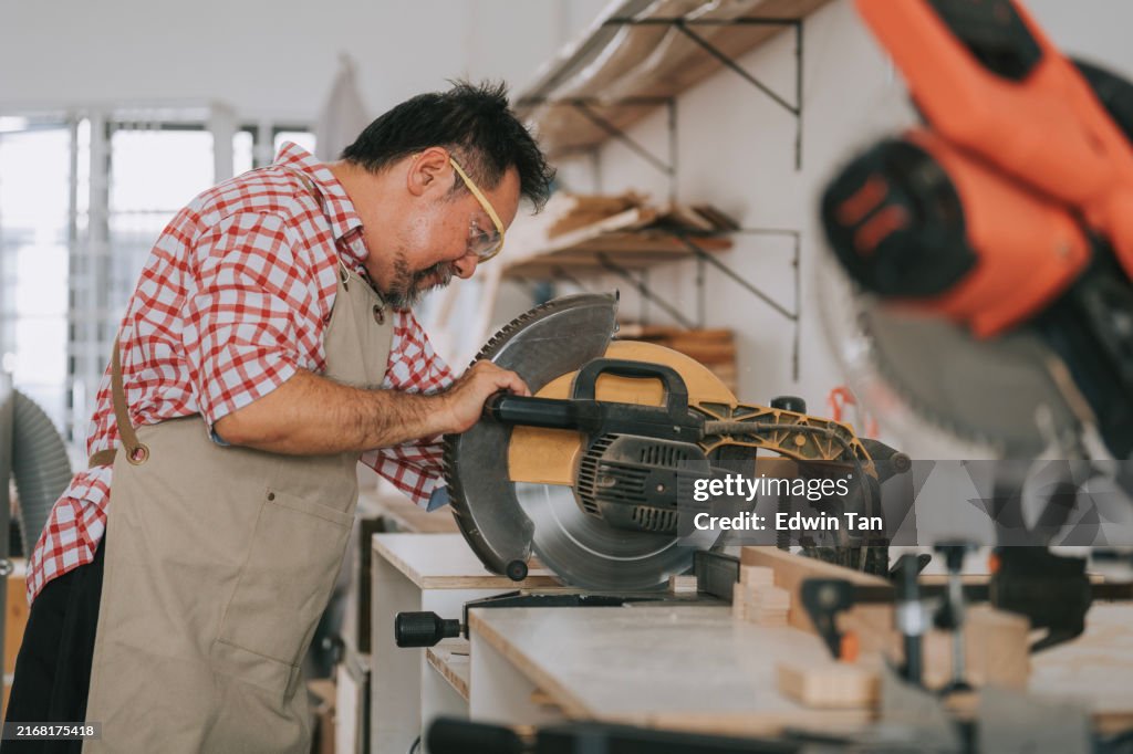 Falegname maschio senior asiatico che taglia il legno con troncatrice in falegnameria officina