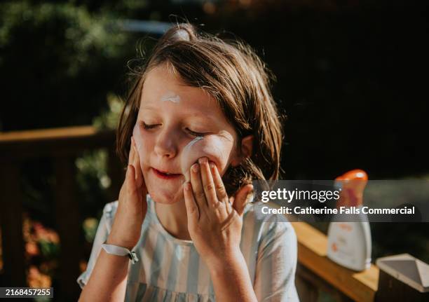 a child is carefully applying sunscreen on her cheeks while standing outside in a backyard. depicts teaching responsibility to children regarding spf and sun protection. - crema de sol fotografías e imágenes de stock