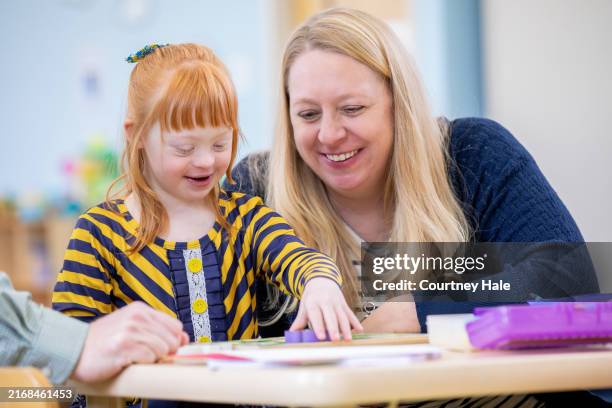 una niña pelirroja y con síndrome de down juega feliz en una pequeña mesa de un salón de clases, con su madre sonriéndole con orgullo. - síndrome de down fotografías e imágenes de stock
