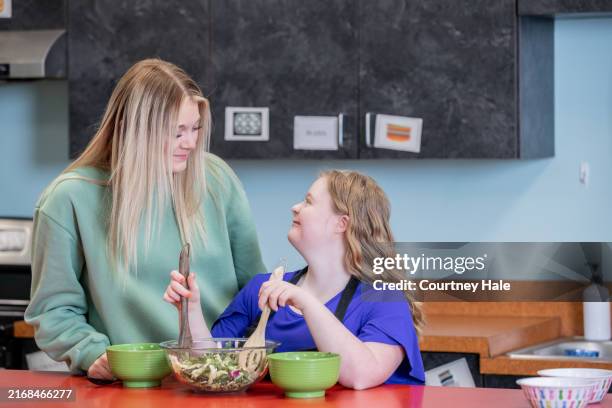 a cheerful teenage girl with down syndrome cooks in the kitchen alongside her sister. - advocacy stock pictures, royalty-free photos & images