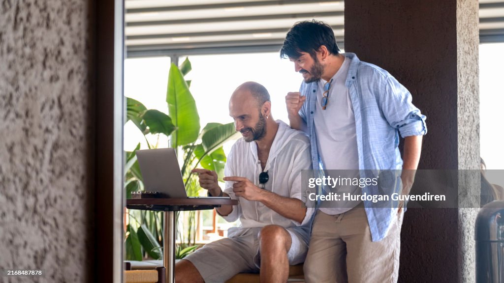 Two middle-aged male professionals are happily working on their laptops in the garden of a café on a summer day. Upon receiving some good news, they celebrate with joyful gestures and expressions