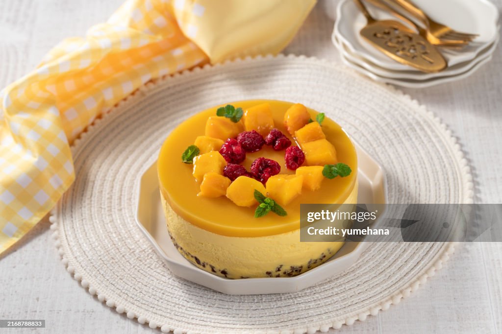 Delicious no bake mango cheesecake with fresh diced mango pulp and raspberry topping on white wooden table.
