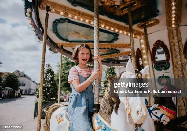 joyful child riding a carousel horse at a fairground on a sunny day, surrounded by trees and festive decorations in the background - carrossel imagens e fotografias de stock