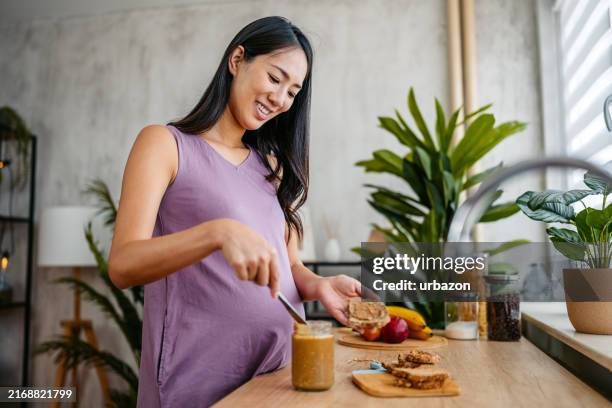 mid-adult pregnant asian woman preparing a peanut butter sandwich in the kitchen at home - peanut butter stock pictures, royalty-free photos & images