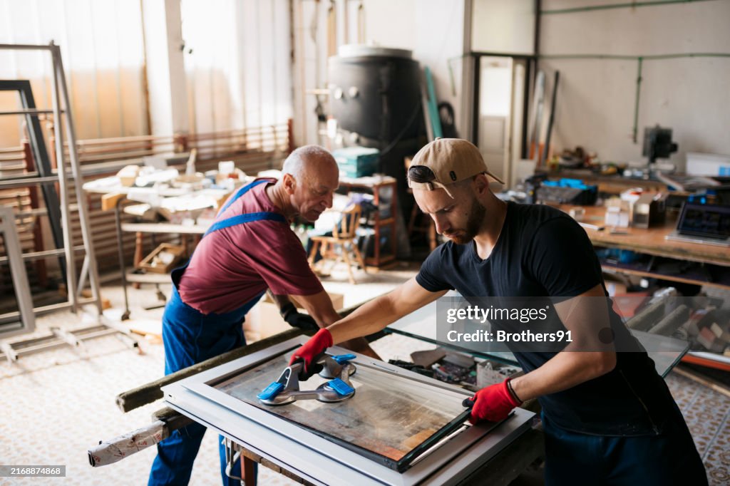 Young worker fitting glass into frame held by senior glazier in workshop