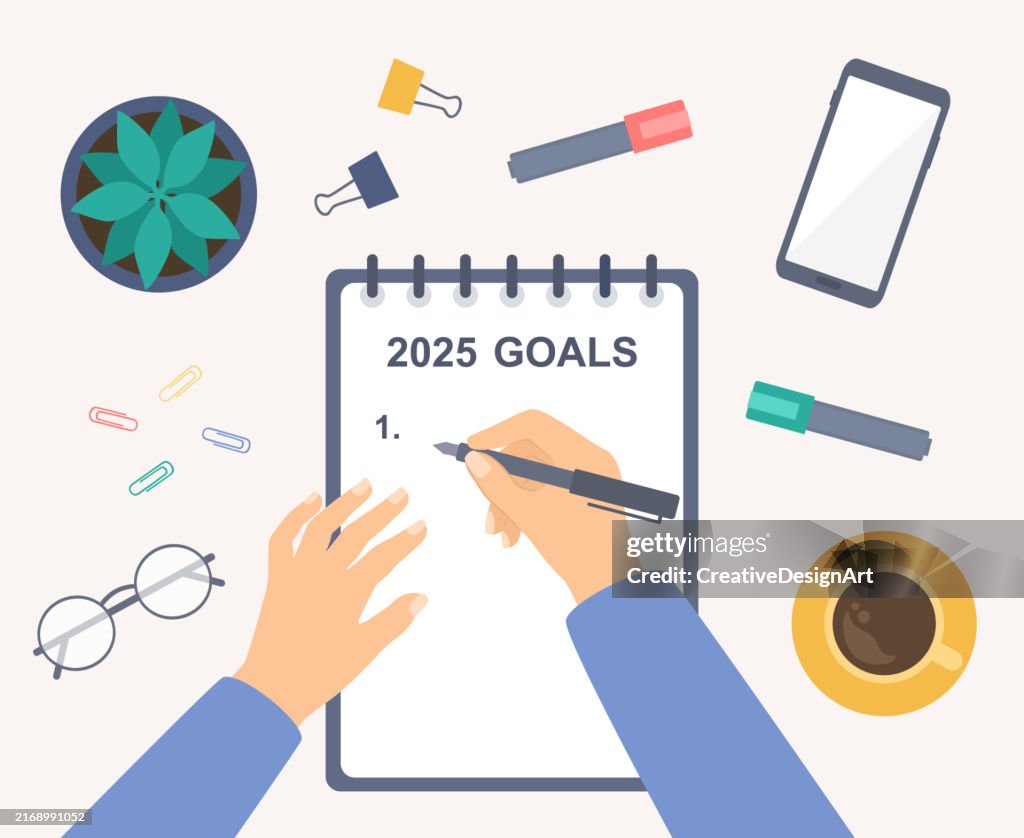 La main tenant un stylo et écrivant les objectifs de 2025 sur un cahier. Vue en plongée de la table avec smartphone, tasse à café, lunettes et plante succulente