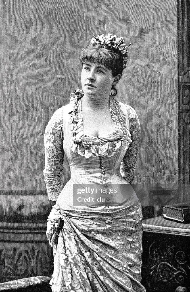 Beauty with a plunging neckline standing , arms on the back, looking up to the side with a flower arrangement in the hair