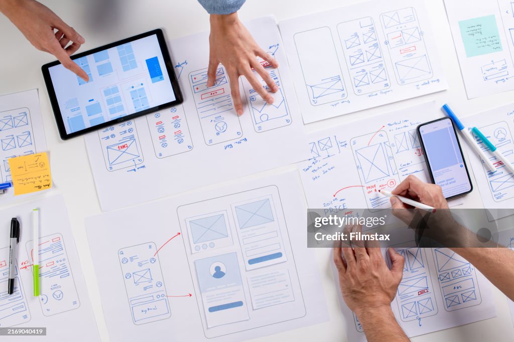 Designers de UX/UI discutindo e brainstorming sobre wireframes para um protótipo de site e aplicativo móvel, cercado por esboços de fluxo de usuário e ferramentas de design, no conceito de design de site e aplicativo móvel.