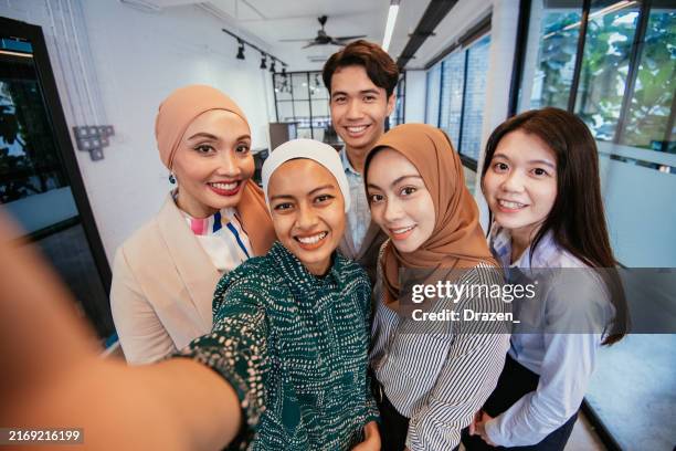 a diverse group of young professionals smiling and taking a selfie together in a modern office environment - indonesische etniciteit stockfoto's en -beelden