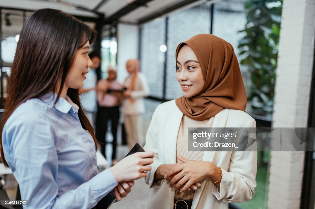 Two Asian businesswomen having a conversation in a modern office environment with colleagues interacting in the background