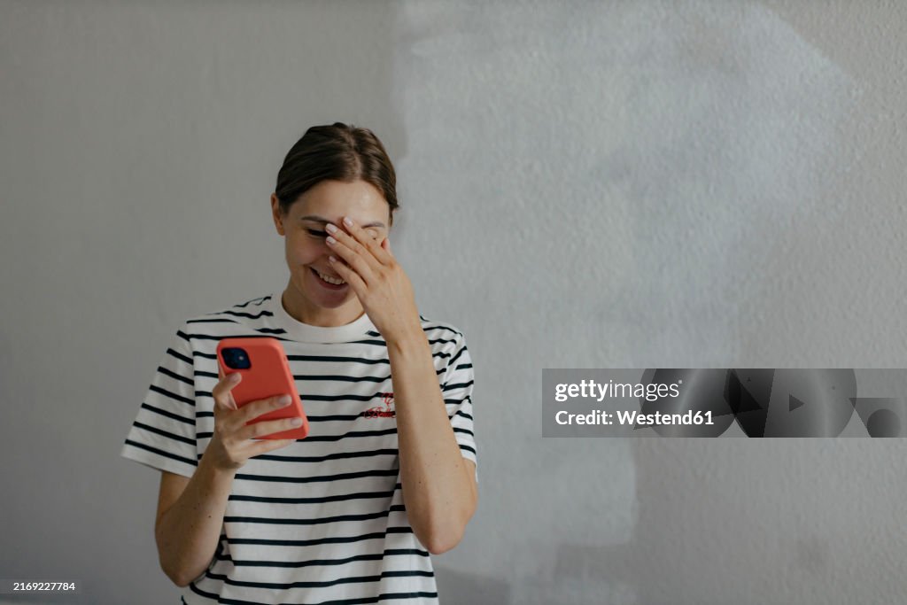 Smiling woman holding smart phone and standing in front of white wall
