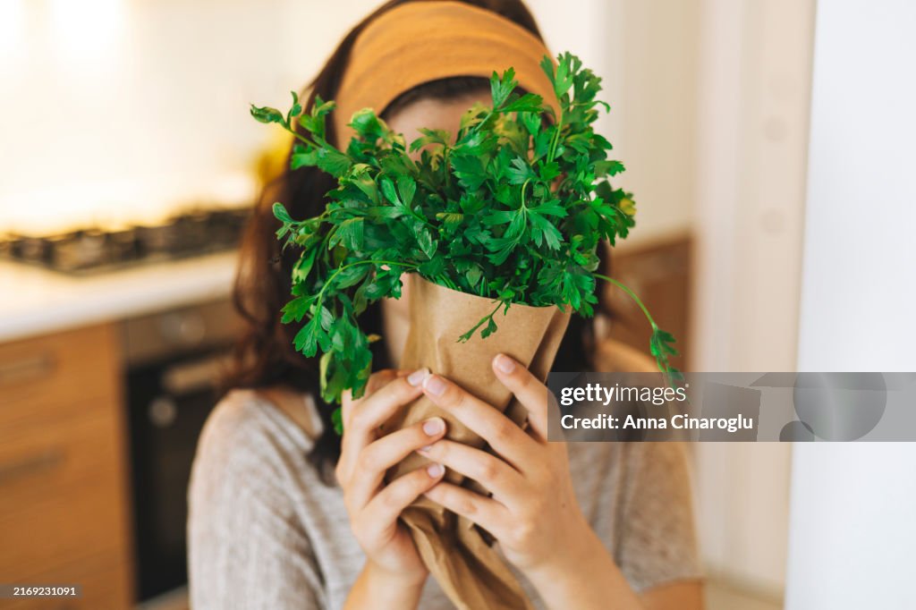 Woman holding fresh parsley while cooking in a modern kitchen with wooden cabinets and a cozy atmosphere during the day