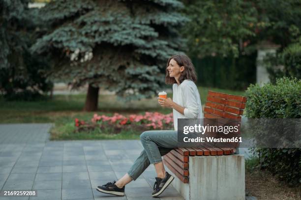 woman sitting on bench holding disposable coffee cup in park - park bench stock pictures, royalty-free photos & images
