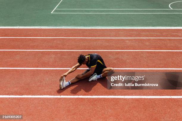 high angle view of male sports person in soccer uniform doing stretching exercises sitting on running track - estadio de atletismo fotografías e imágenes de stock