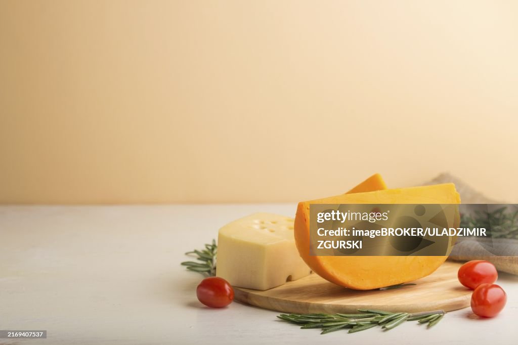 Various types of cheese with rosemary and tomatoes on wooden board on a white and orange background and linen textile. Side view, close up, copy space, selective focus