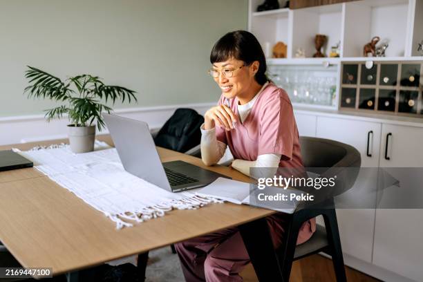 fröhliche krankenschwester mit laptop bei der arbeit von zu hause aus - medizinerkleidung stock-fotos und bilder