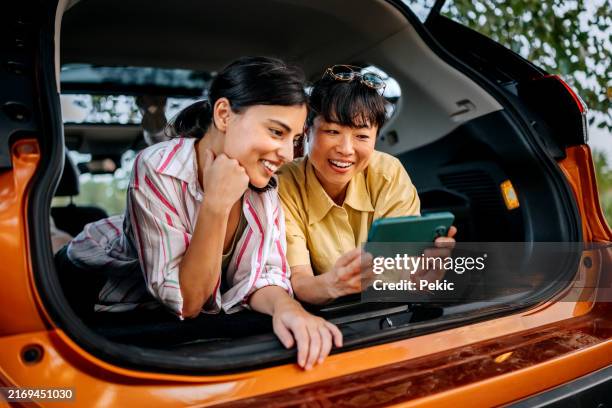 two smiling female friends using smart phone while laying in car trunk together - allongé sur le devant photos et images de collection
