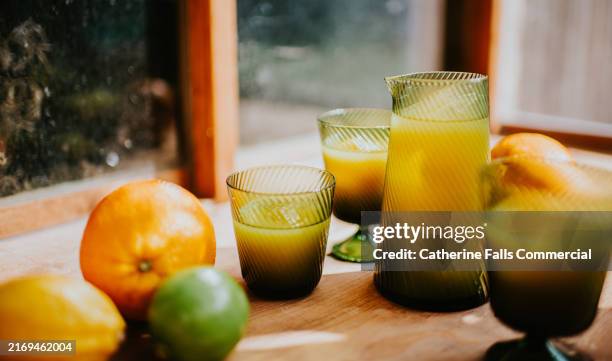a small jug of orange juice and fresh citrus juices with fruits on a wooden table in natural light during a sunny morning - orange juice stock pictures, royalty-free photos & images